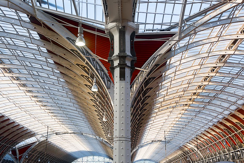A detailed interior view of a modern architectural structure featuring a large, curved, ribbed metal and glass ceiling supported by a prominent grey riveted steel column at the center. The ceiling has multiple layers with a combination of transparent and opaque panels, allowing natural light to illuminate the space below. Several metal pendant lights hang from the ceiling, enhancing the brightly lit environment. The structure's design incorporates red accents visible behind the steel framework, suggesting the interior of a transport hub or shopping arcade. This setting highlights elements typical of the interiors involved in home relocation and furniture transport processes, where structural details and lighting are crucial for planning removal routes and assessing space for loading furniture and packing materials. The image reflects an environment associated with busy moving logistics, supported by a professional removals company such as Removal Company Paddington.