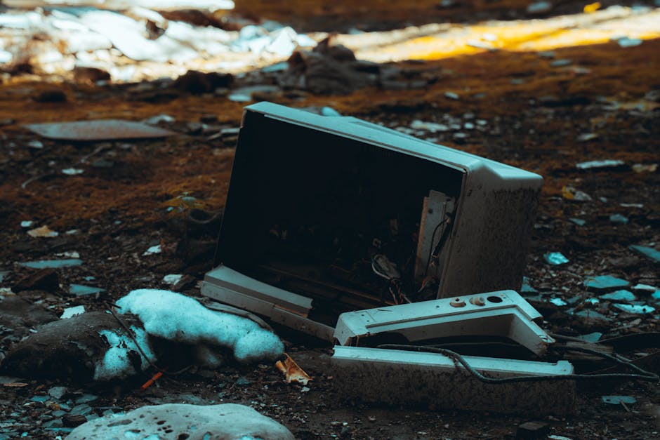 A discarded, broken microwave oven lying on the ground outdoors, surrounded by patches of snow and scattered debris, including broken plastic and paper fragments. The microwave's door is open, exposin