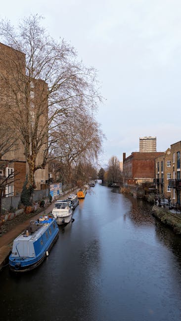 A view of the Little Venice canalside in W9 showing narrow waterway lined with leafless trees and residential buildings, including brick and stone structures, on both sides. Several boats are moored a