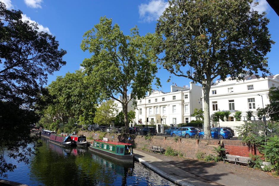A view of the Little Venice canalside in W9 showing narrow waterway lined with leafless trees and residential buildings, including brick and stone structures, on both sides. Several boats are moored a