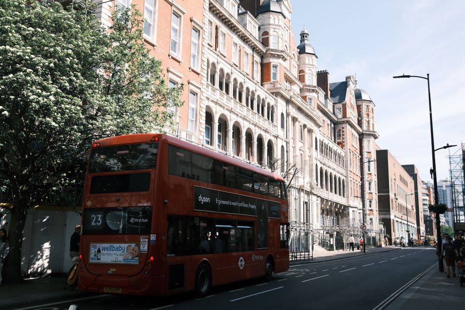 A red double-decker bus numbered 23 is parked on a street in Paddington during daytime, with ornate Victorian-style buildings featuring turrets, arches, and decorative brickwork lining the background.