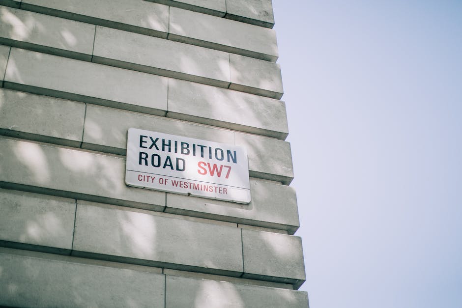 A close-up photograph of a street sign mounted on the exterior corner of a building made of light grey stone bricks. The sign reads 'Exhibition Road SW7' in bold black and red letters, indicating the