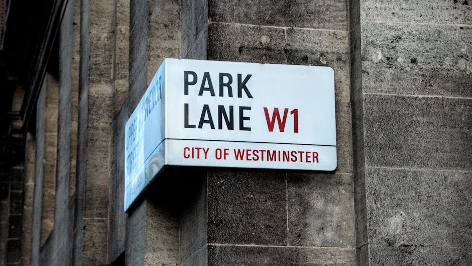 A close-up photograph of a street sign mounted on the exterior corner of a building made of light grey stone bricks. The sign reads 'Exhibition Road SW7' in bold black and red letters, indicating the